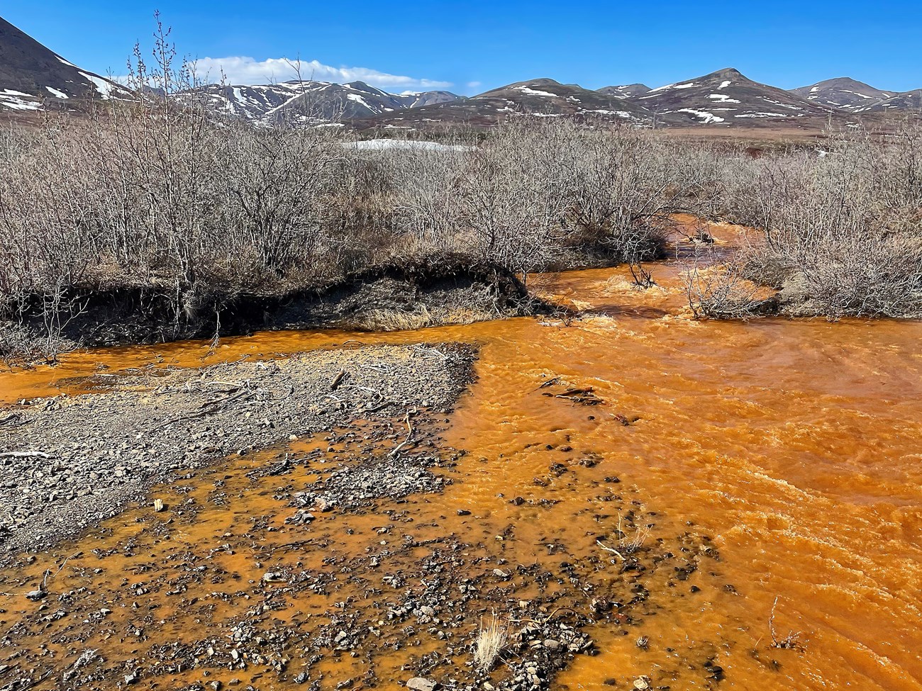 Rushing stream where all of the water is bright orange. Mountains covered in patches of snow line the horizon.