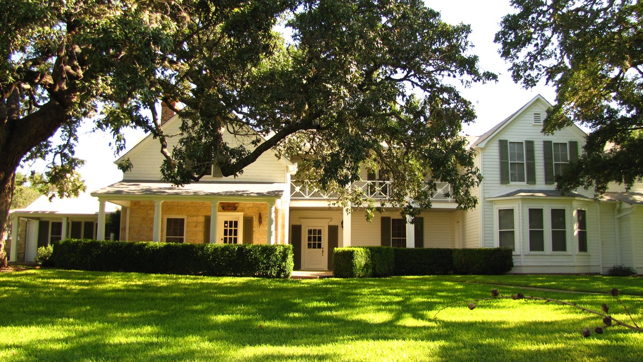 A two-story white frame and stone house is shaded by a large tree.