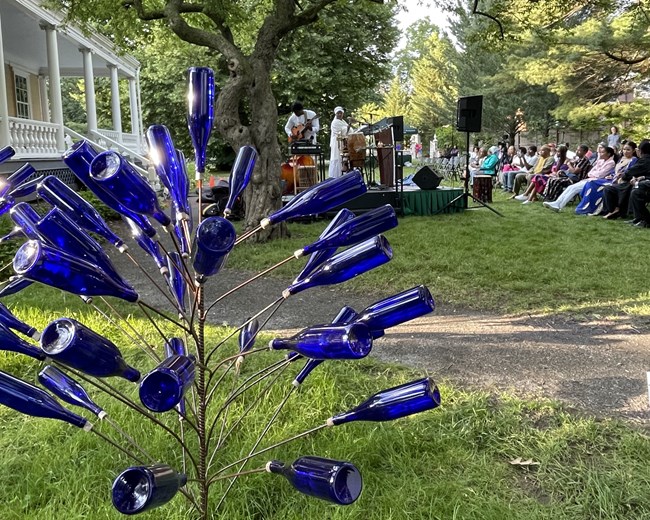 Crowd outdoors listening to music and poetry with blue glass bottle tree in foreground.