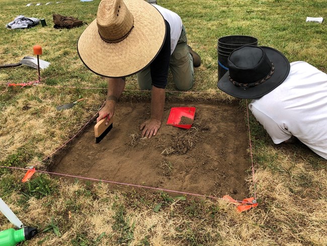 Two students work with brushes and trowels on a square excavation unit.