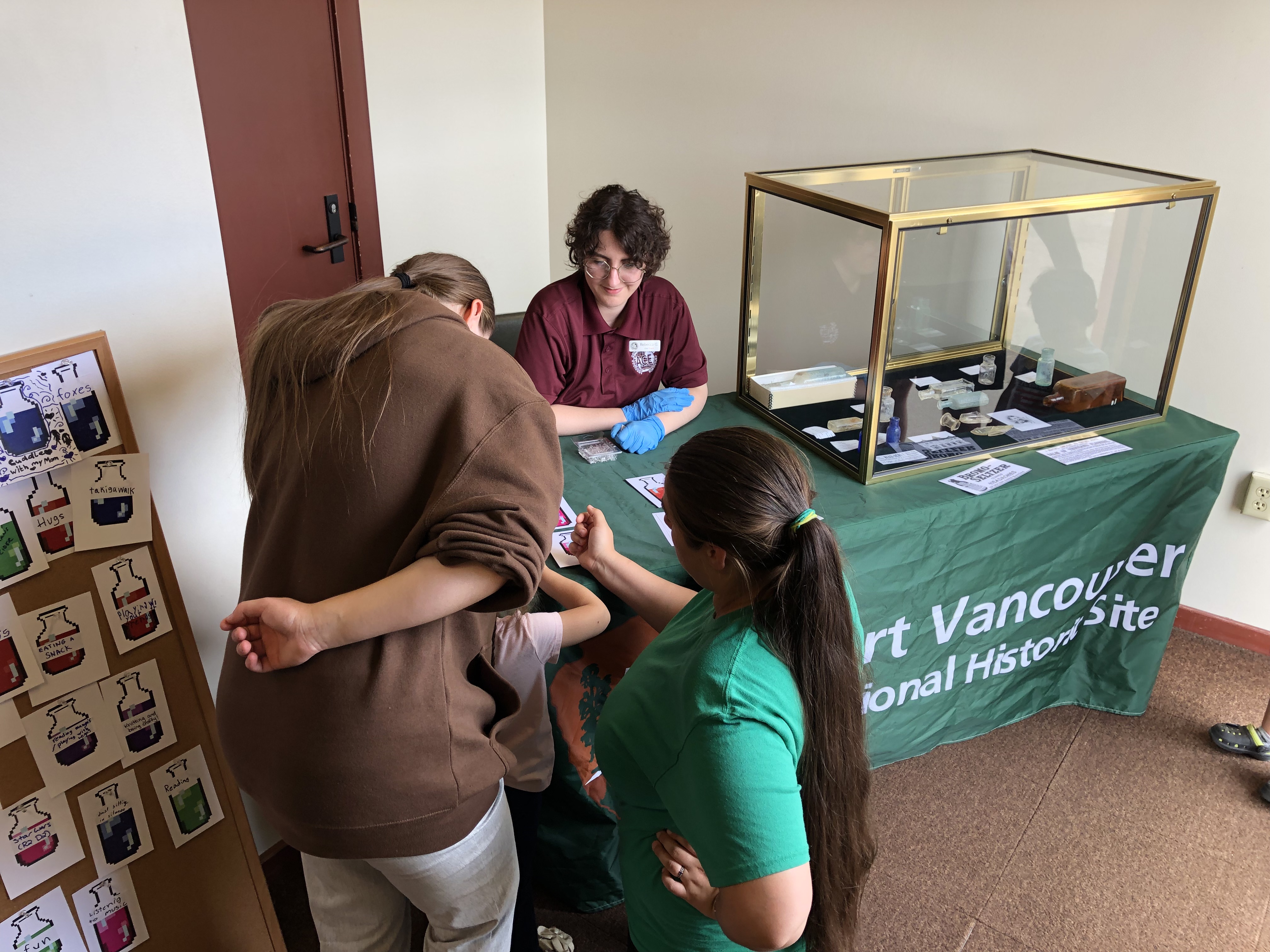 A person sits at a table next to an exhibit case with archaeological artifacts while visitors work on an activity.