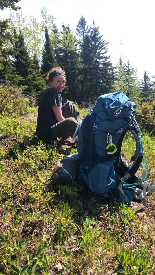 A woman sits on the ground next to a large backpack in a grassy forest.