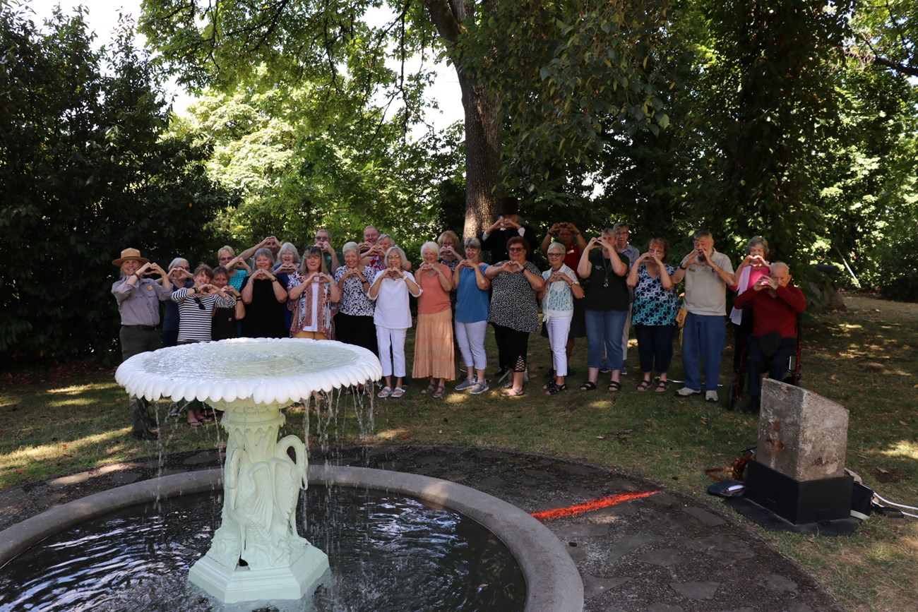 A group of people, including one person in a National Park Service uniform, stand behind the fountain holding their hands up to make a heart shape.