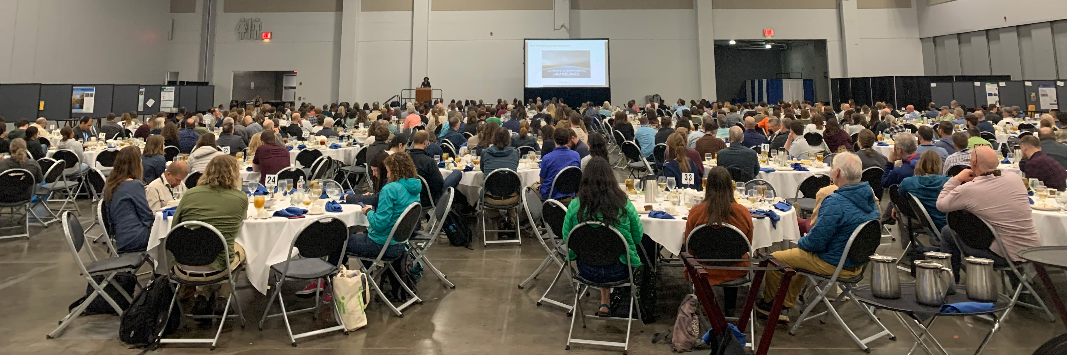 Large room full of people sitting at round tables with a podium and large screen in the background.