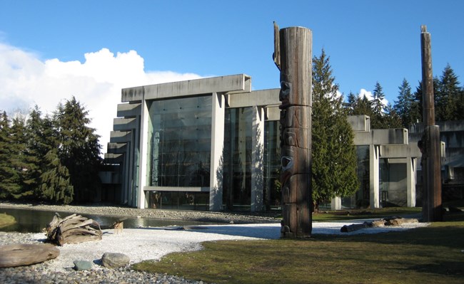 A large steel and glass building with totems, woods, and a water feature.
