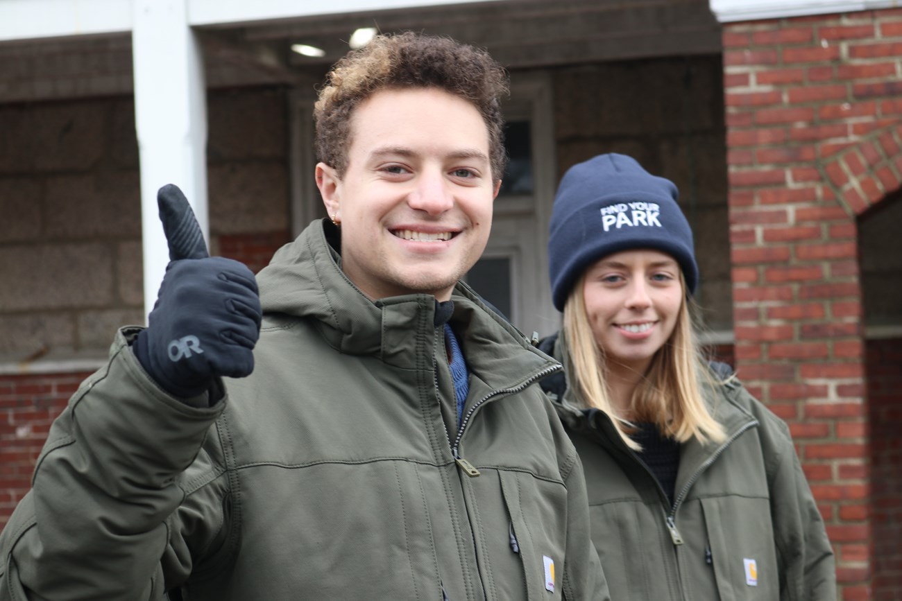 Two Corps Members smile in front of a brick building; one corp members gives a thumb's up.