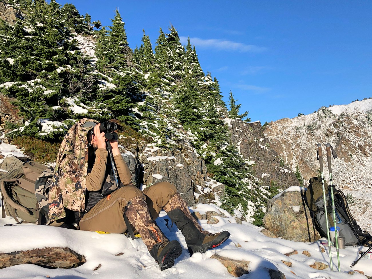 Person in camouflaged winter gear, sitting in the snow, looking through binoculars.