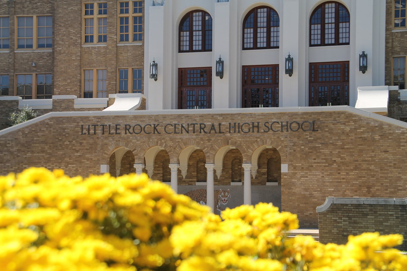The front entrance of Little Rock Central High School, a historic brick building with arched windows and columns, seen behind bright yellow flowers in the foreground.