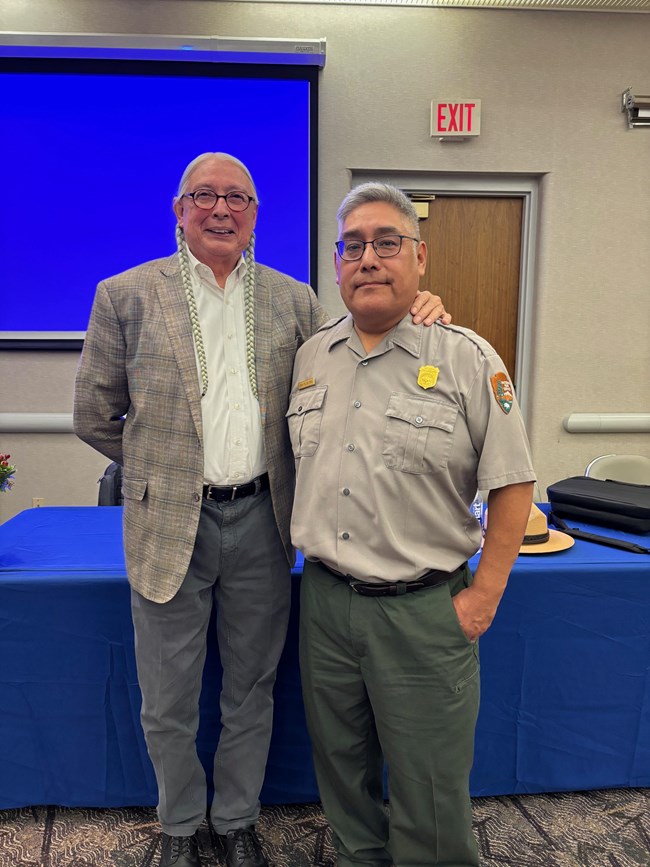 A man in a park ranger uniform standing with a man with braided hair
