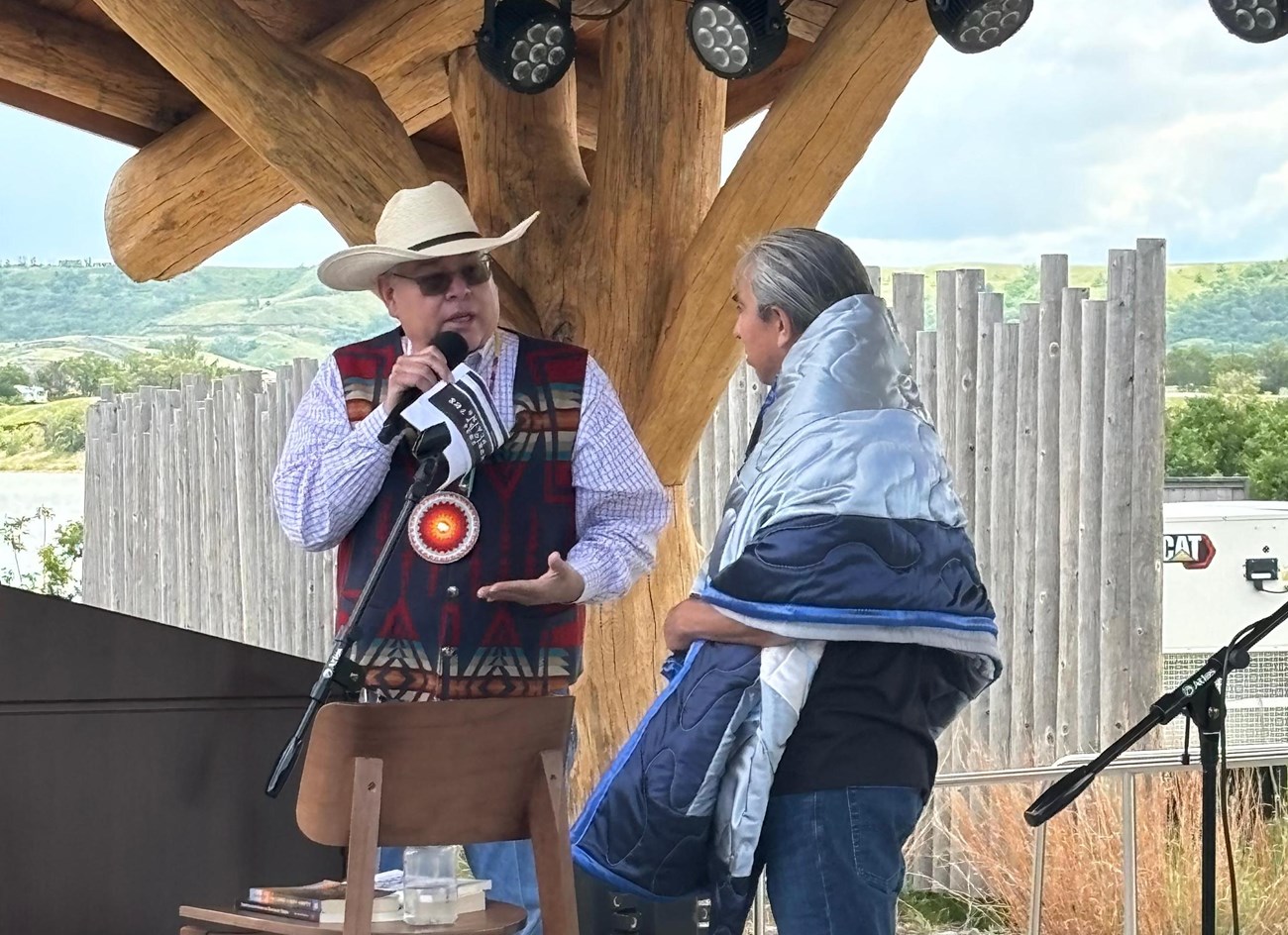 A man wrapped in a blanket looking at a man speaking into a microphone