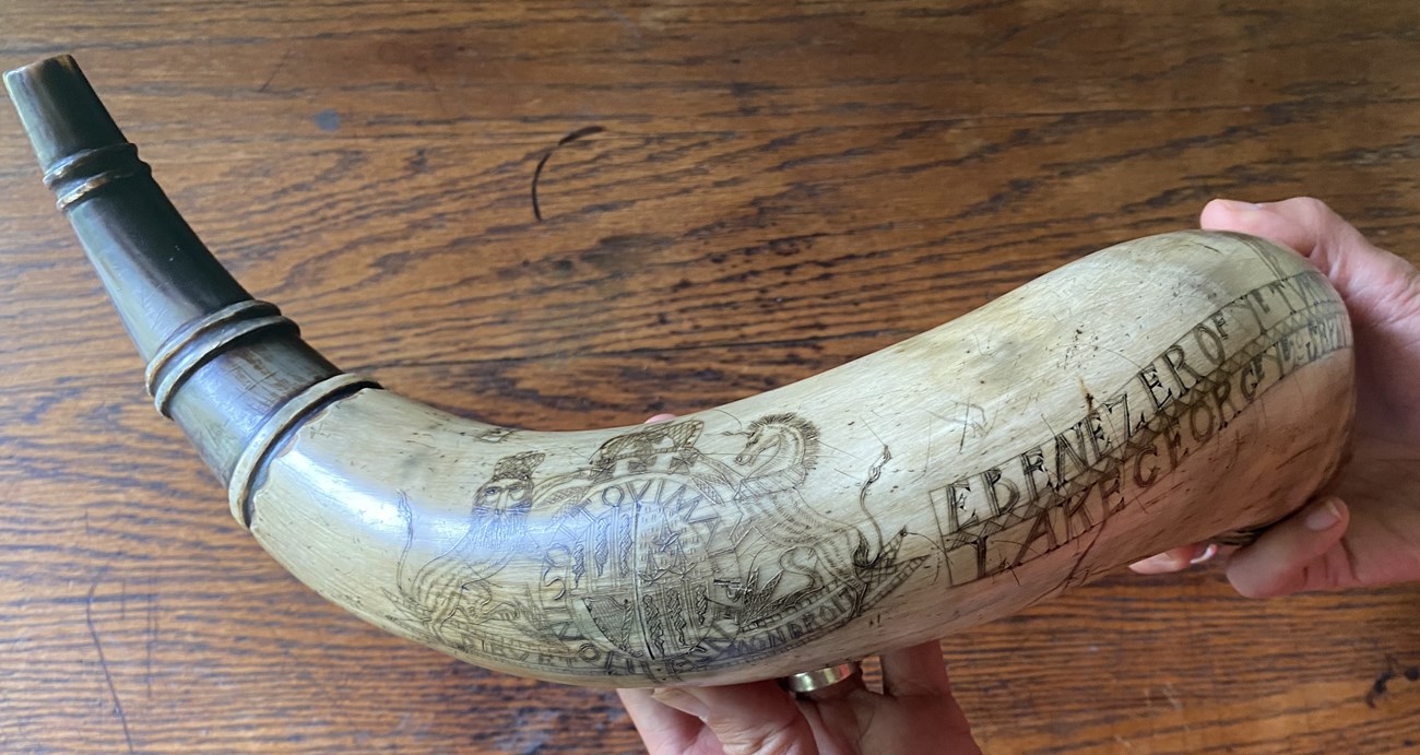 A large powder horn made of cow bone, with images of the British Royal Coat of Arms and name Ebenezer scratched into the surface