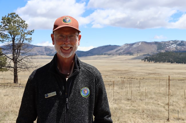 A man with a white beard and glasses wears a NPS volunteer jacket. He stands in a montane grassland and smiles at us.