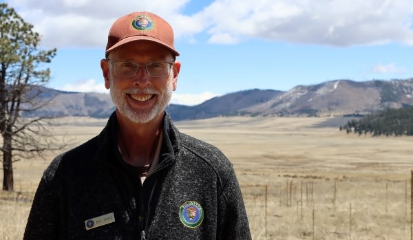 A portrait of a smiling man wearing a brown ballcap and a black fleece jacket standing outdoors.