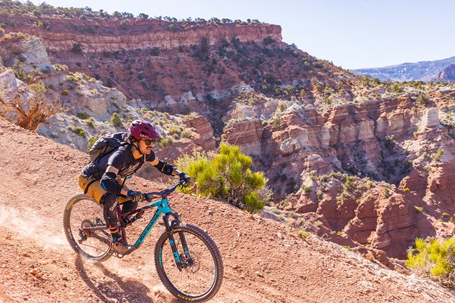 A man on a mountain bike rides down a red dirt trail. A colorfully striped canyon wall rises in the background.