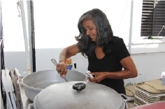 Woman with black and grey shoulder-length hair stands in an outdoor kitchen stirring a large pot of rice.