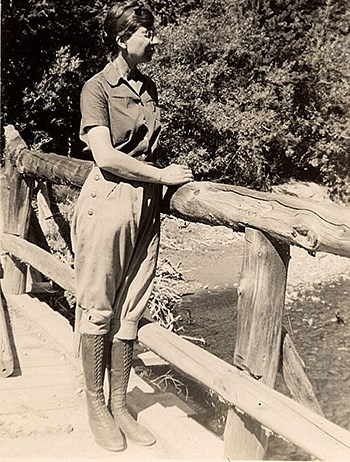 Isabel Bassett Wasson wearing breeches, boots, and a blouse stands on a bridge, looking into the distance.