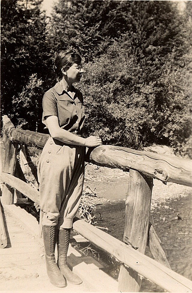 Isabel Wasson standing on a wooden bridge wearing breeches, knee-high boots, and a short-sleeved shirt.