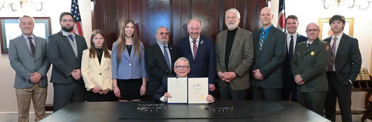 A group of people stand behind a table that a man sits at, holding a sheaf of papers.