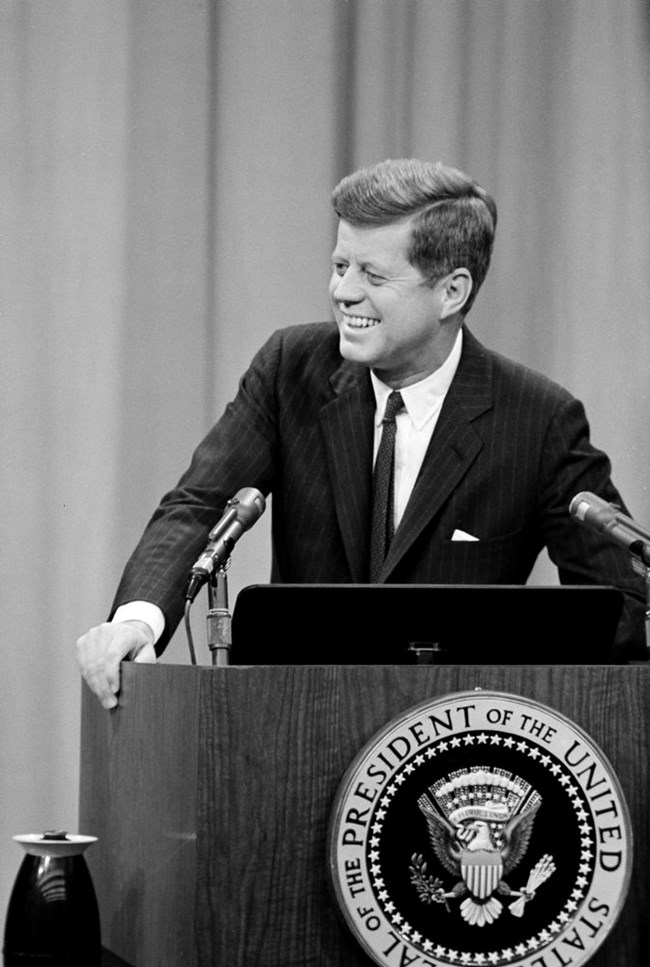 President Kennedy laughs as he stands behind a podium with the presidential seal
