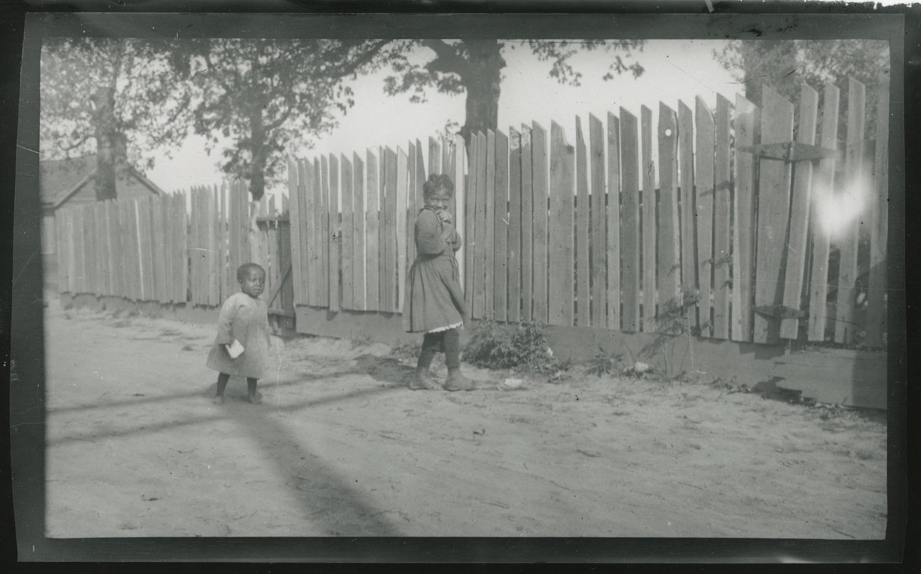 Two young children stand on a dirt road by a wood fence.