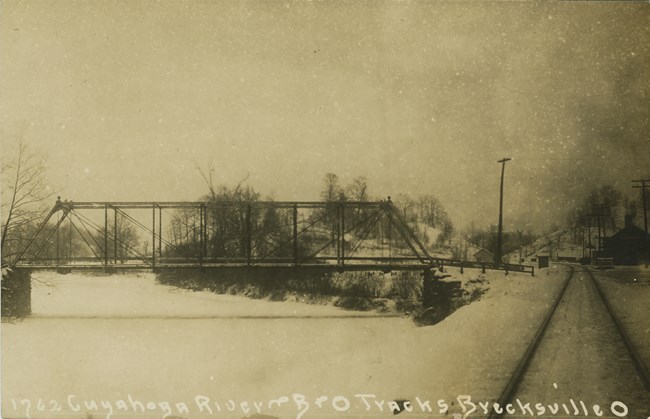 Side view of a metal truss bridge over a frozen river blanketed in snow with railroad tracks and small buildings, right.