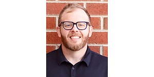 Headshot of Jorgenson in front of a brick wall. He's smiling, with a short beard, moustache, and short, light hair, wearing black glasses and a black, short-sleeved collared shirt.