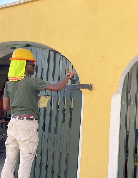 A worker with a yellow hardhat holds a paint brush and pan as they paint a grey gate along a yellow wall.