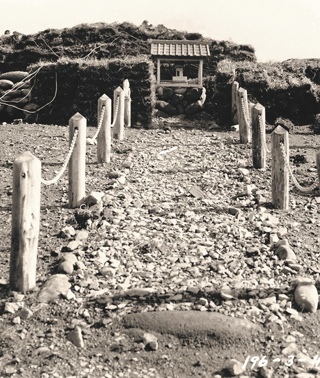black and white photo of pathway lined with rope and posts to a small shrine, with sandbags on both sides.