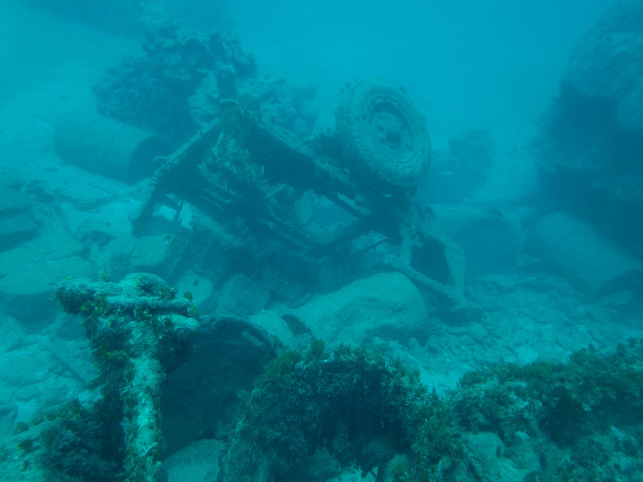 Underwater image of a rusty, algae-covered vehicle wreck, surrounded by scattered debris and rocks.