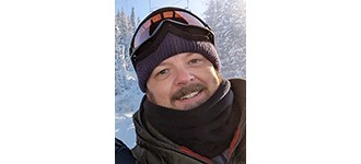 Lonneker outside in a warm coat, ski hat, and with ski goggles on the of his hat, smiling for the camera.