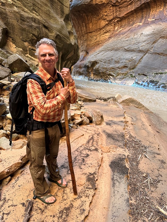 A man hiking through Zion National Park stands with a wooden walking stick and backpack on a sandy riverbank, surrounded by towering red and tan sandstone canyon walls.