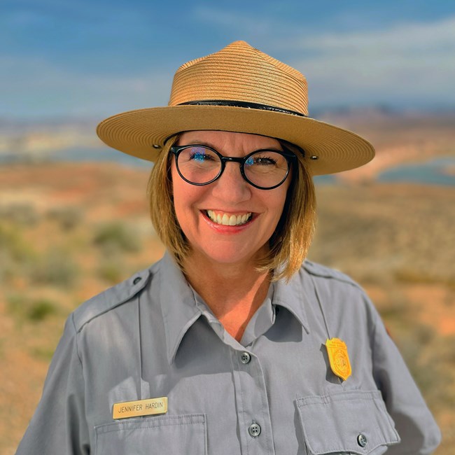 Smiling park ranger wearing a flat-brimmed ranger hat and a gray uniform shirt smiling and standing outdoors under blue skies