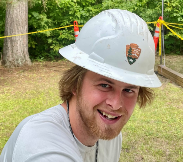 Worker smiles into the camera while wearing a white hardhat with national park service Arrowhead emblem on the front.