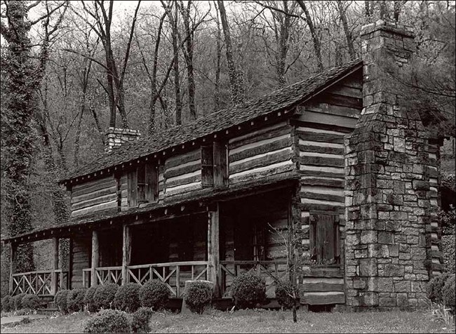 2-story wood house with porch, 3 windows, and 2 stone chimneys. House sits in front of a dense forest.