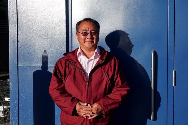 an alaska native man smiling and wearing a red jacket against the backdrop of a blue building.
