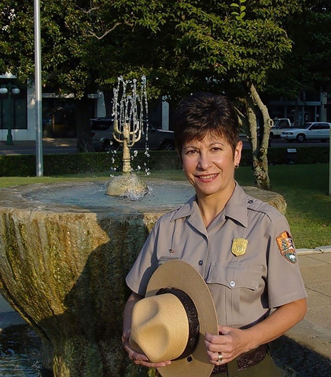 Josie Fernandez poses with her NPS uniform with flat hat in her hand, standing in front of a hot spring.