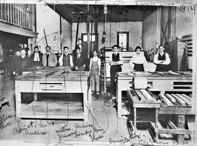 Group of 11 people standing in a room with exposed brick walls and concrete floors surrounded by newspaper printing machinery
