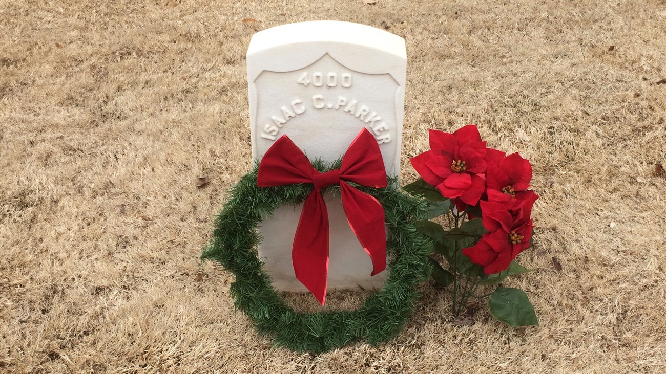 A white headstone in brown grass with a wreath and poinsettia plant laid beside it. Text reads, "4000, Isaac C. Parker."