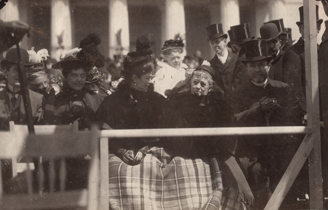 Black and white photo of two women with hats and a blanket covering their laps while sitting among a crowd