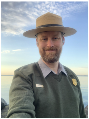 Photo of Justin Olson in his ranger uniform smiling outdoors with a lake in the background.