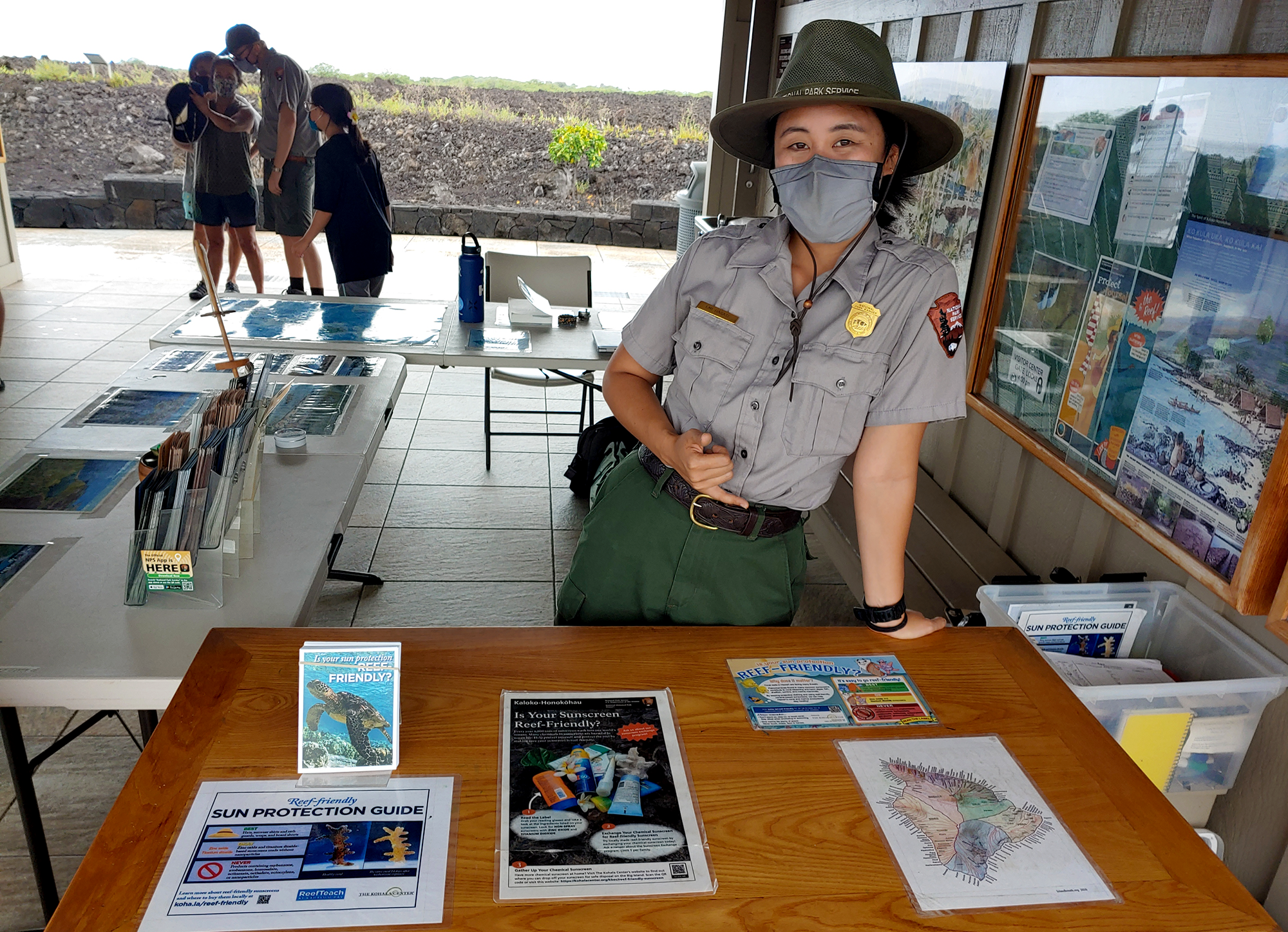 A woman in an NPS uniform standing behind a table with sunscreen information on it. In the background is another ranger and three visitors.