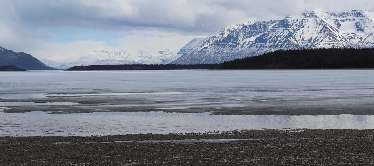 A cover of snow highlights the contours of tall mountains, rising into clouds on the opposite shore of a lake.