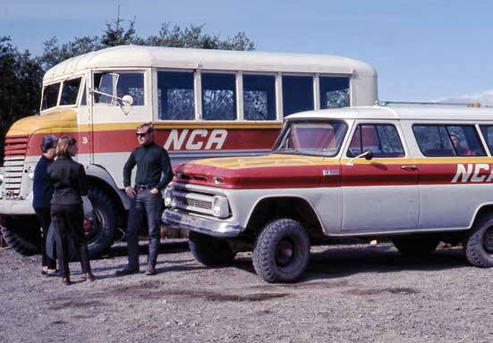 Three people stand near the front of a jeep and a bus, both painted cream with a red and yellow stripe.