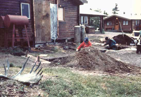 A person kneels beside a hole and dirt pile behind a log structure during excavation work.