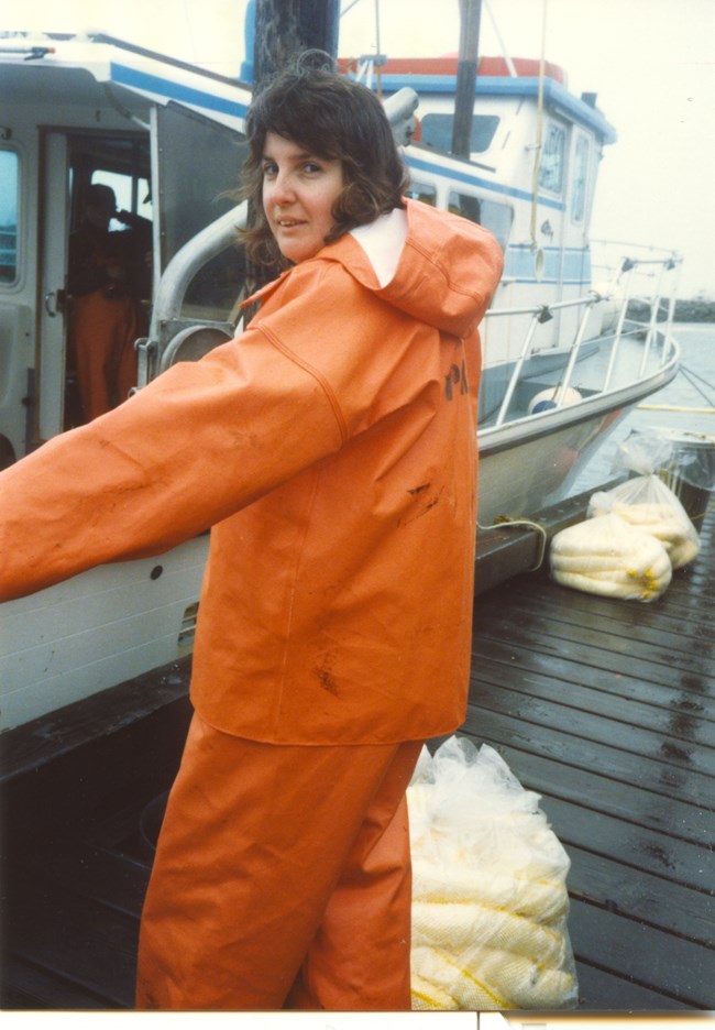 Woman in orange rain gear next to clean-up supplies and boat on dock