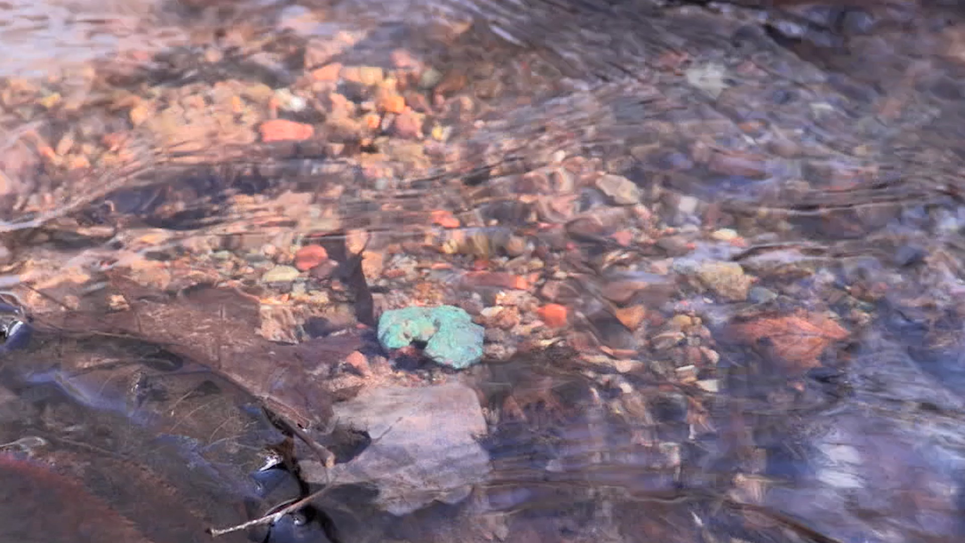 Closeup photo of a gentle stream of crystal clear water flowing over pebbles including a piece of float copper with a light green patina.