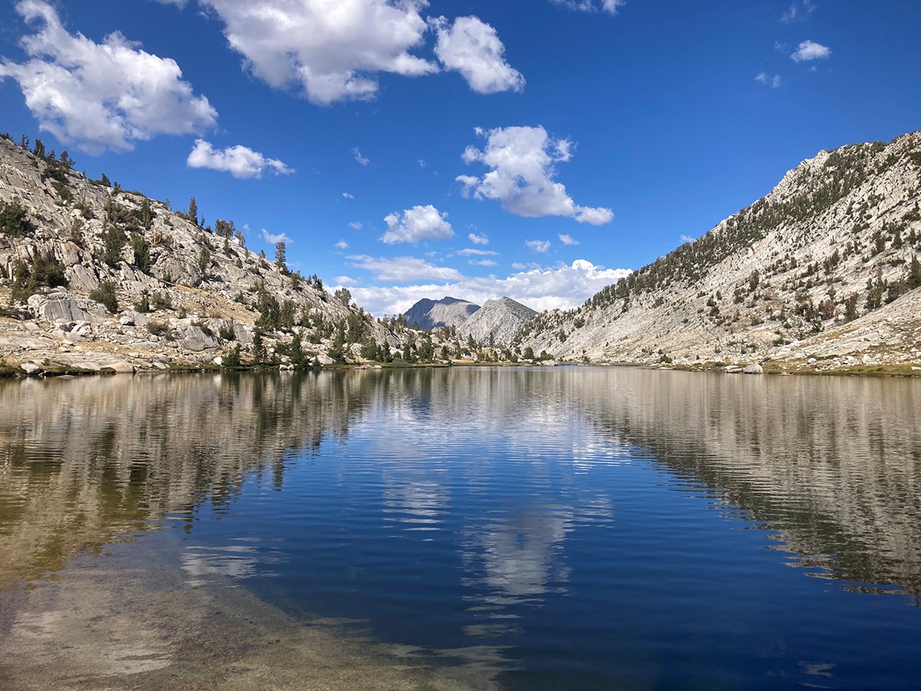 View of a large lake with clear water reflecting sky, clouds, and mountains.