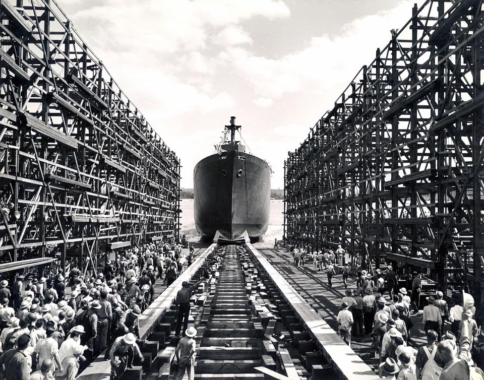 A black and white photo of a ship launching into the Columbia River. A crowd of workers stand with their backs to the camera, watching the ship slide into the water.