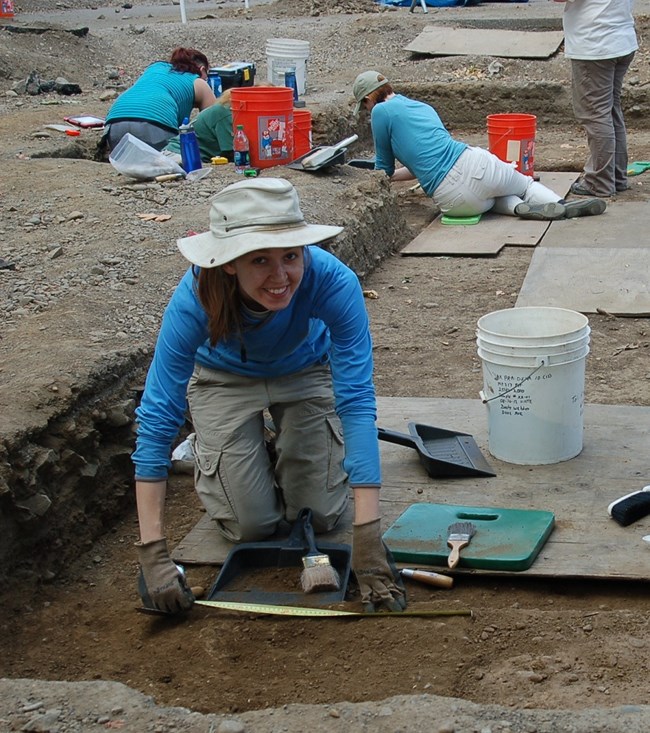 A woman kneels down to use a tape measure to measure an excavation unit.