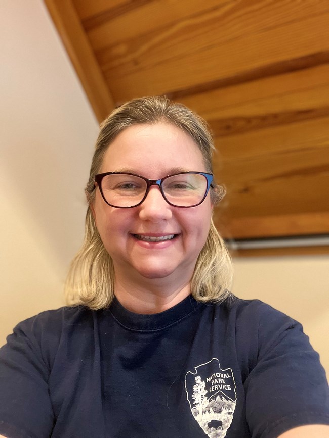 a blonde woman wearing glasses and a t-shirt with the NPS arrowhead on the chest smiles for the camera